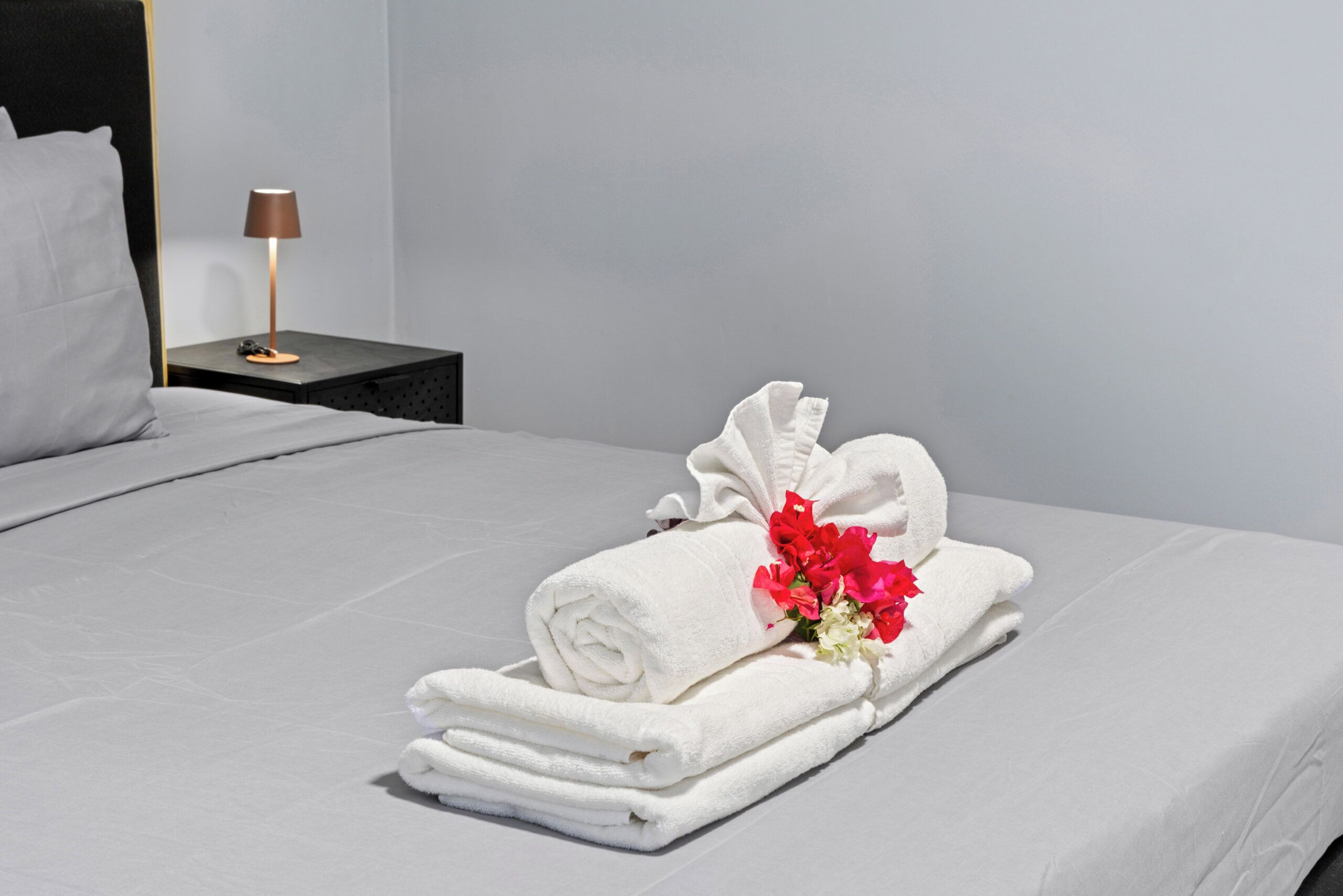 Un lit bien fait, avec du linge de lit blanc et une serviette enroulée ornée de fleurs rouges, dans une chambre d’hôtel.