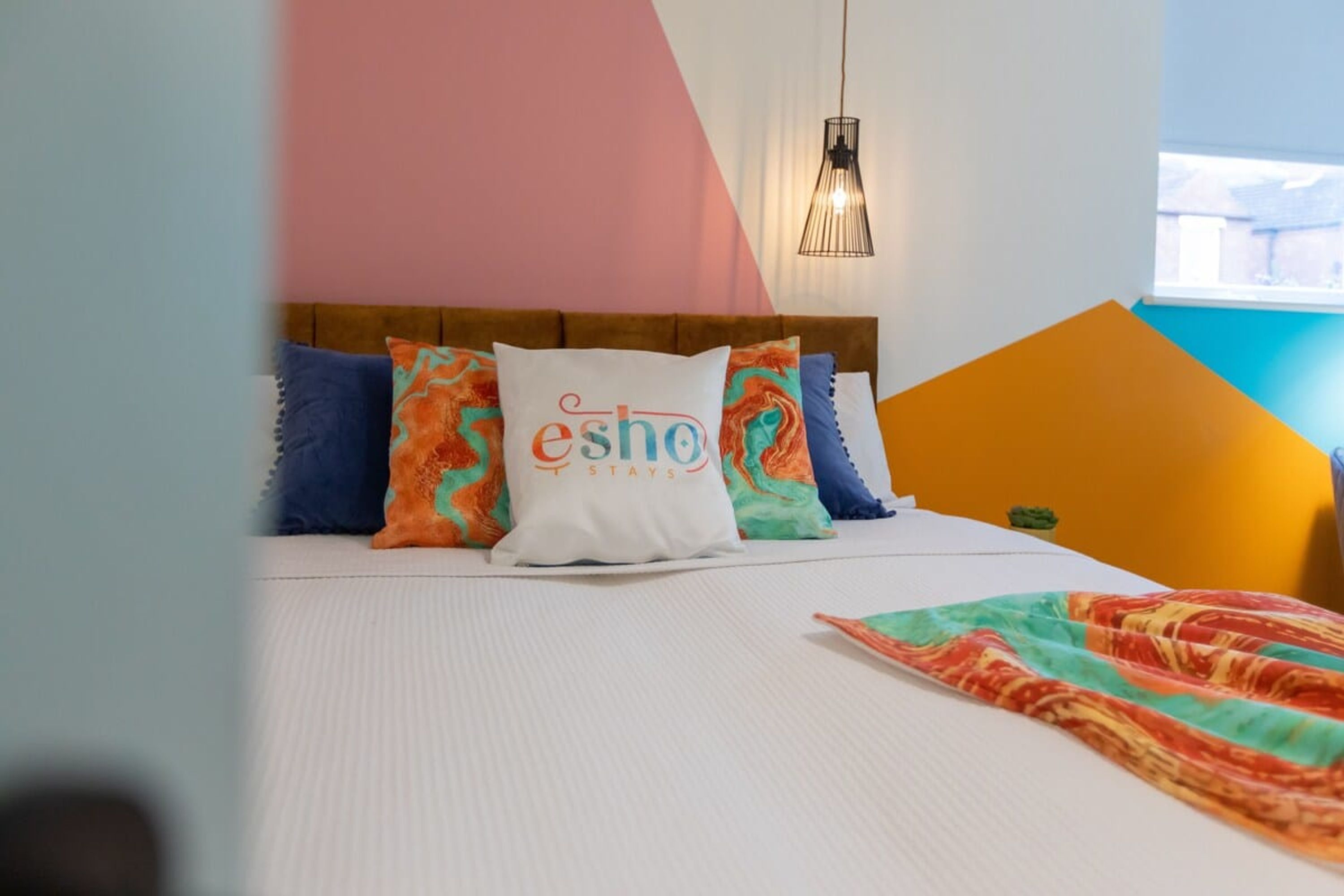 Un letto con cuscini colorati e una coperta, una testiera in legno e una lampada a sospensione.
