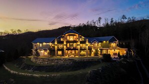 A large, multi-story house with a stone facade, multiple windows, and a well-lit exterior. - Bear Necessities Lodge: 16,000 SqFt Estate | Indoor Pool, Pickleball, Theater!!! (Sevierville)
