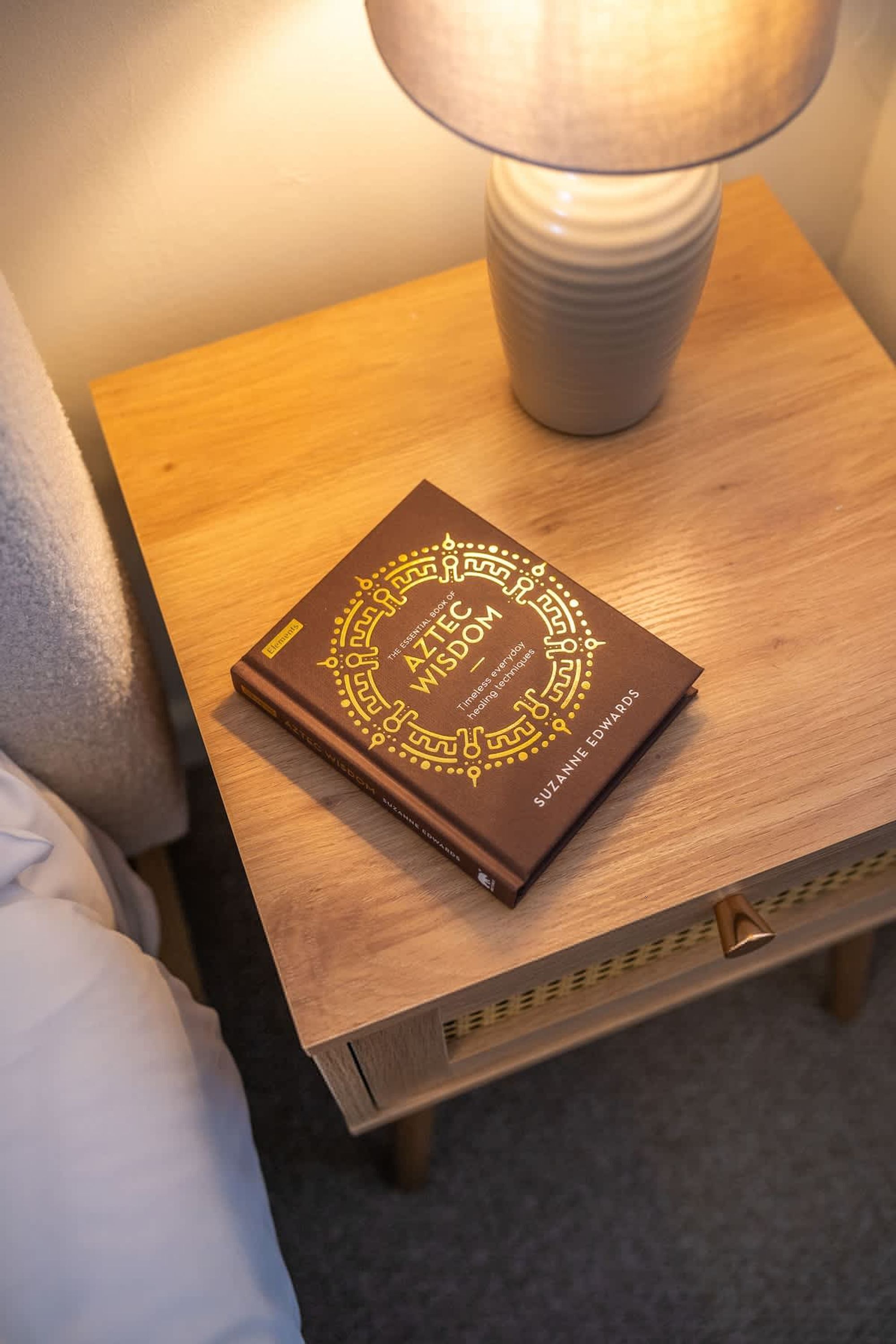 Un livre intitulé « Sagesse aztèque » est posé sur une table de chevet en bois, à côté d’une lampe.