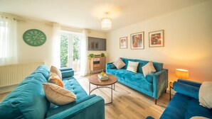 A living room with a blue sofa, a round coffee table, and a clock on the wall. - 5 BD 3 Bath 3 Car Drive Large Garden Pet Friendly (Cirencester)