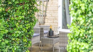 An outdoor seating area with wicker furniture, a table with a bottle and glasses, and a brick wall in the background. - 5 BD 3 Bath 3 Car Drive Large Garden Pet Friendly (Cirencester)