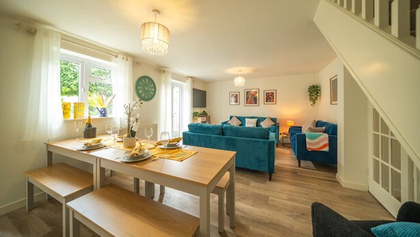 A dining area with a wooden table and benches, a teal sofa, and a staircase. - 5 BD 3 Bath 3 Car Drive Large Garden Pet Friendly (Cirencester)