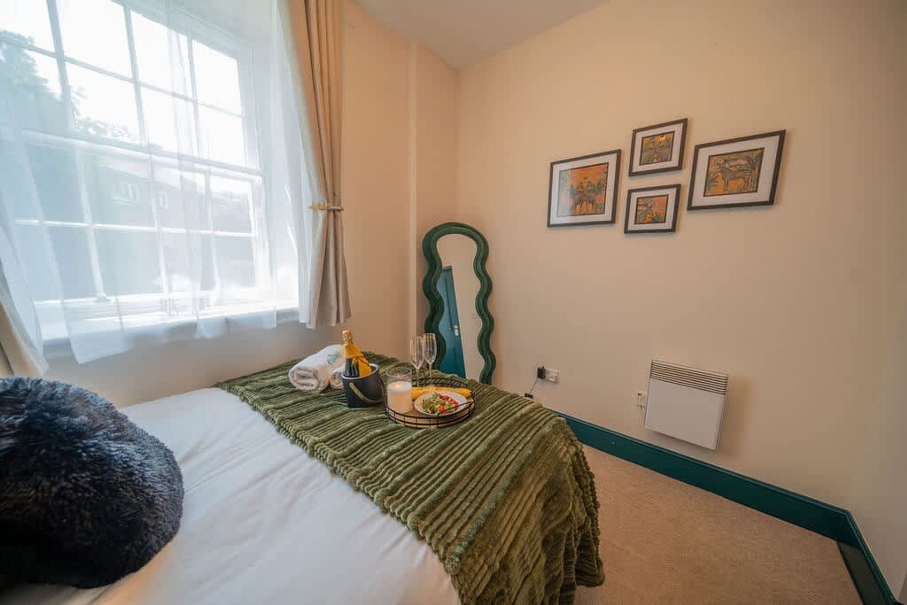 Ein Hotelzimmer mit Bett, einem Fenster mit Vorhängen, einem grünen Kopfteil, einem fest installierten Heizkörper und gerahmten Bildern.