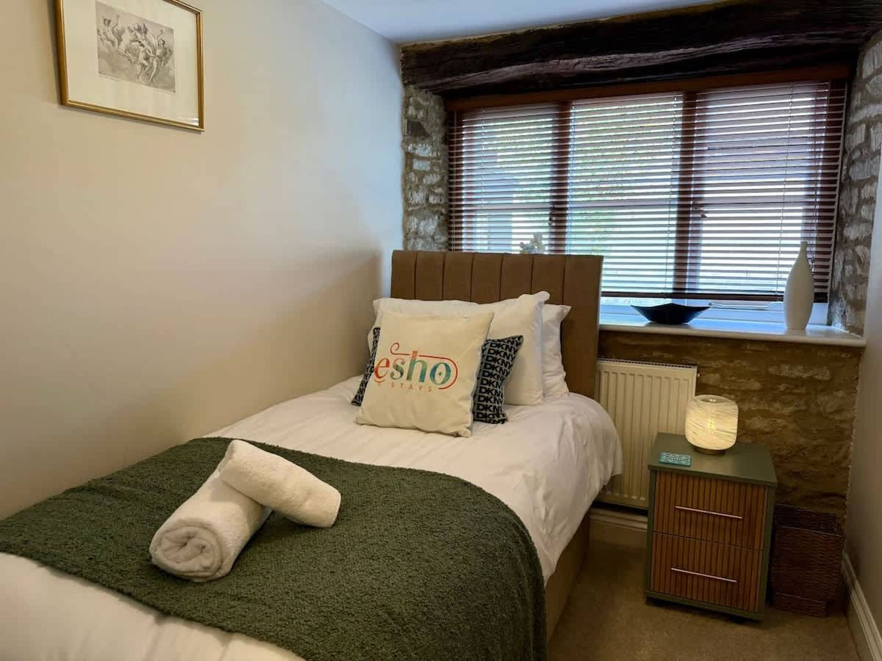 A neatly made bed with a green blanket, a pillow with the text 'esho stays,' and a framed picture on the wall.