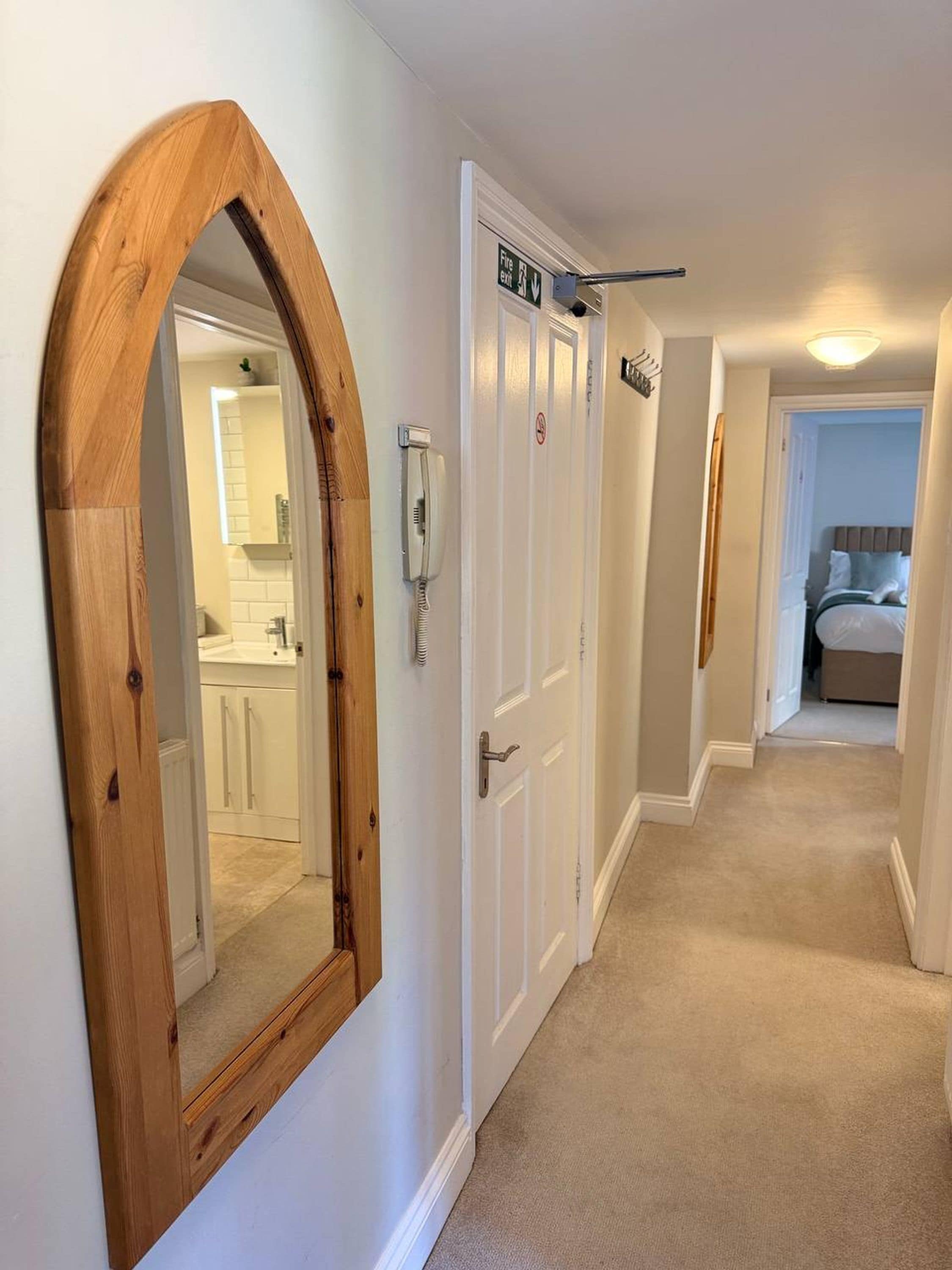 Un couloir avec un miroir encadré en bois, des portes blanches, un panneau indiquant la sortie de secours, un téléphone et un lit visible au fond.