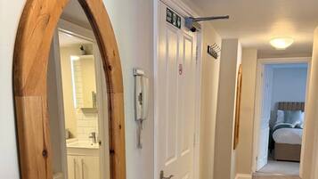 Un couloir avec un miroir encadré en bois, des portes blanches, un panneau indiquant la sortie de secours, un téléphone et un lit visible au fond.