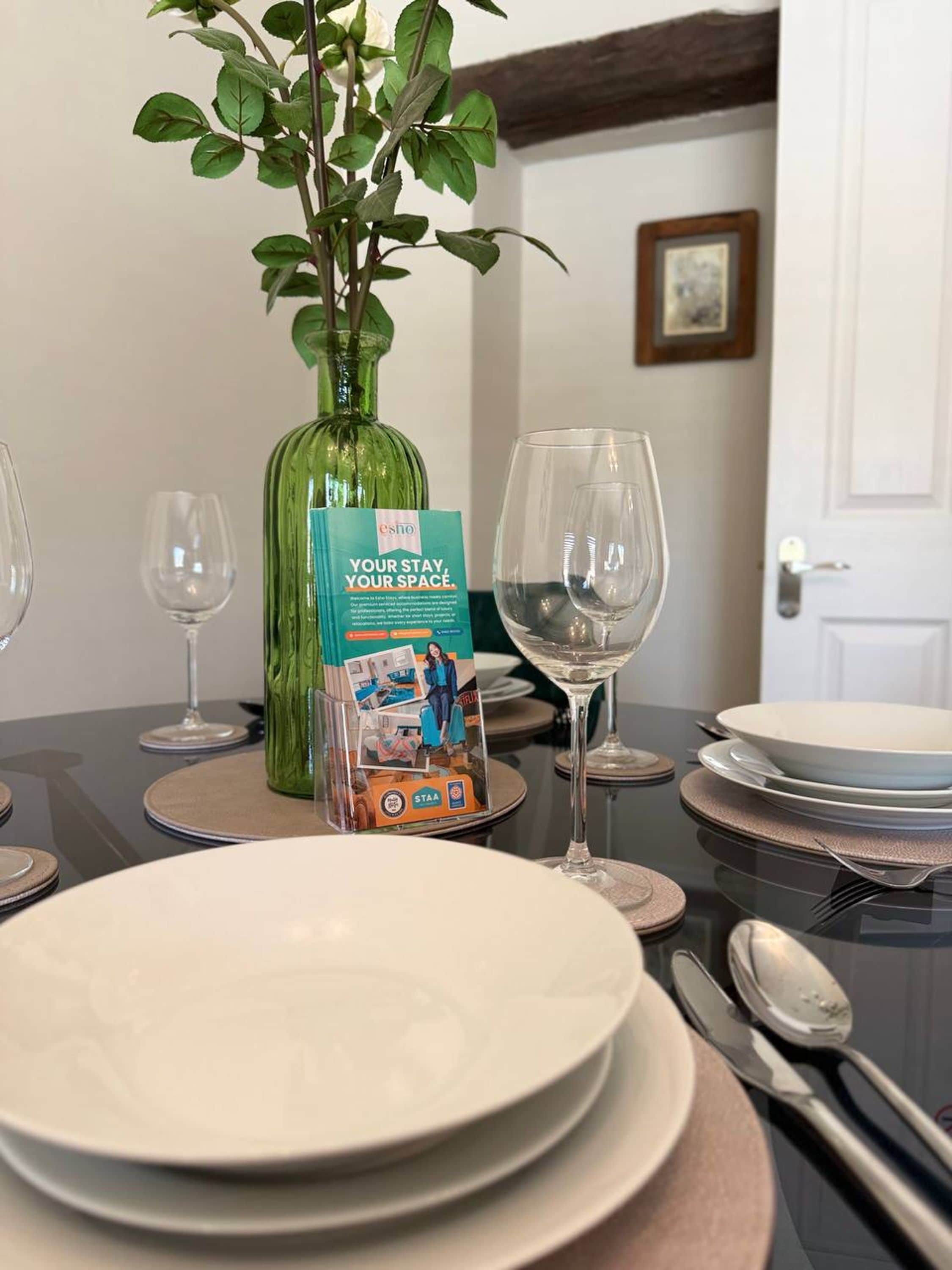 Una mesa de comedor servida con platos, copas de vino y cubiertos, un jarrón verde con una planta, y un folleto titulado 'Your Stay, Your Space'.