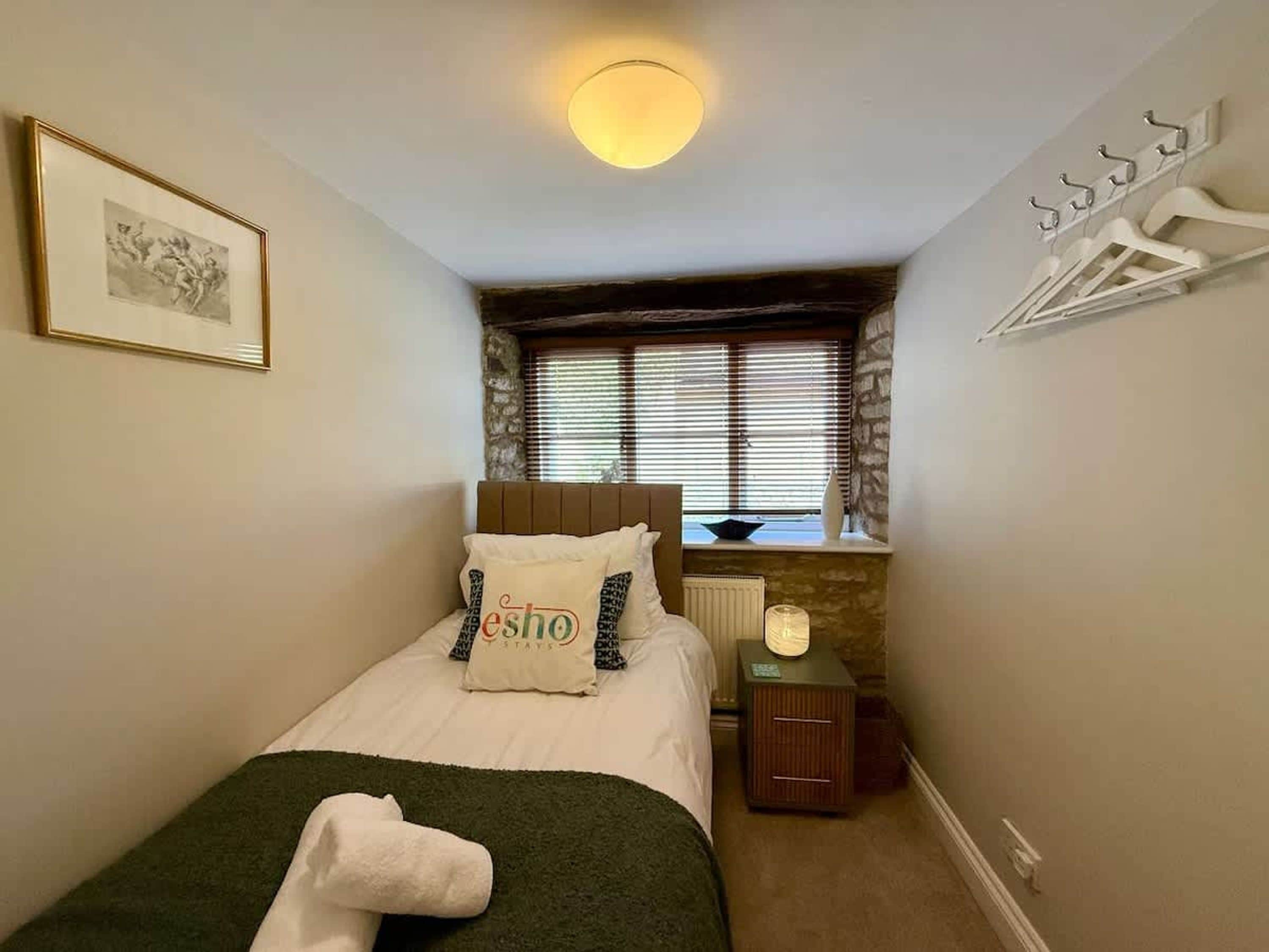 Quarto com cama, mesa de cabeceira, radiador, janela com persianas, cabideiro fixado na parede e um quadro emoldurado.