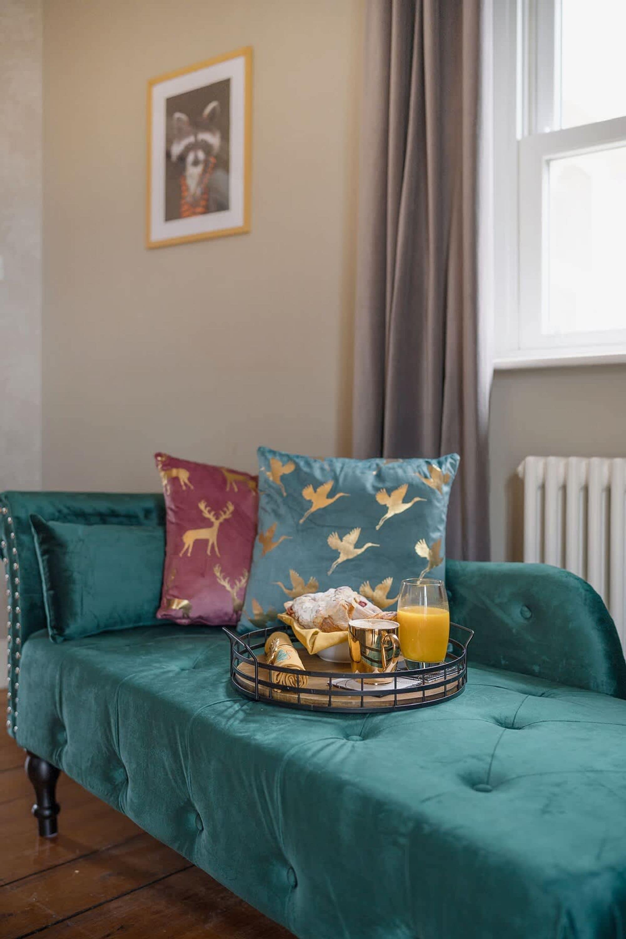 A teal sofa with decorative pillows, a tray with breakfast items, and a window with curtains.