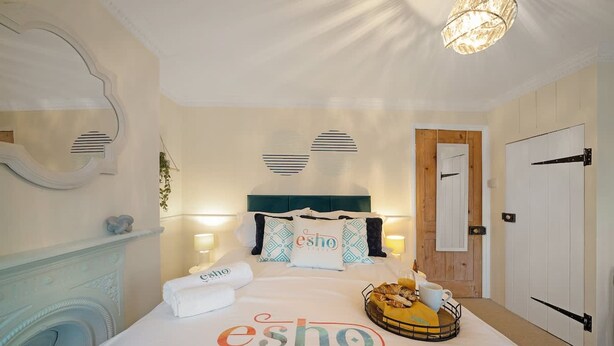 A hotel room with a bed, a headboard, a bedside table, a lamp, a mirror, and a door. - Cotswolds 3BD 1 5 Bath Garden City Centre (Cirencester)
