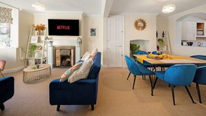 A living room with a fireplace, a TV mounted on the wall, a dining area with blue chairs, and a kitchen visible through an archway. - Cotswolds 3BD 1 5 Bath Garden City Centre (Cirencester)