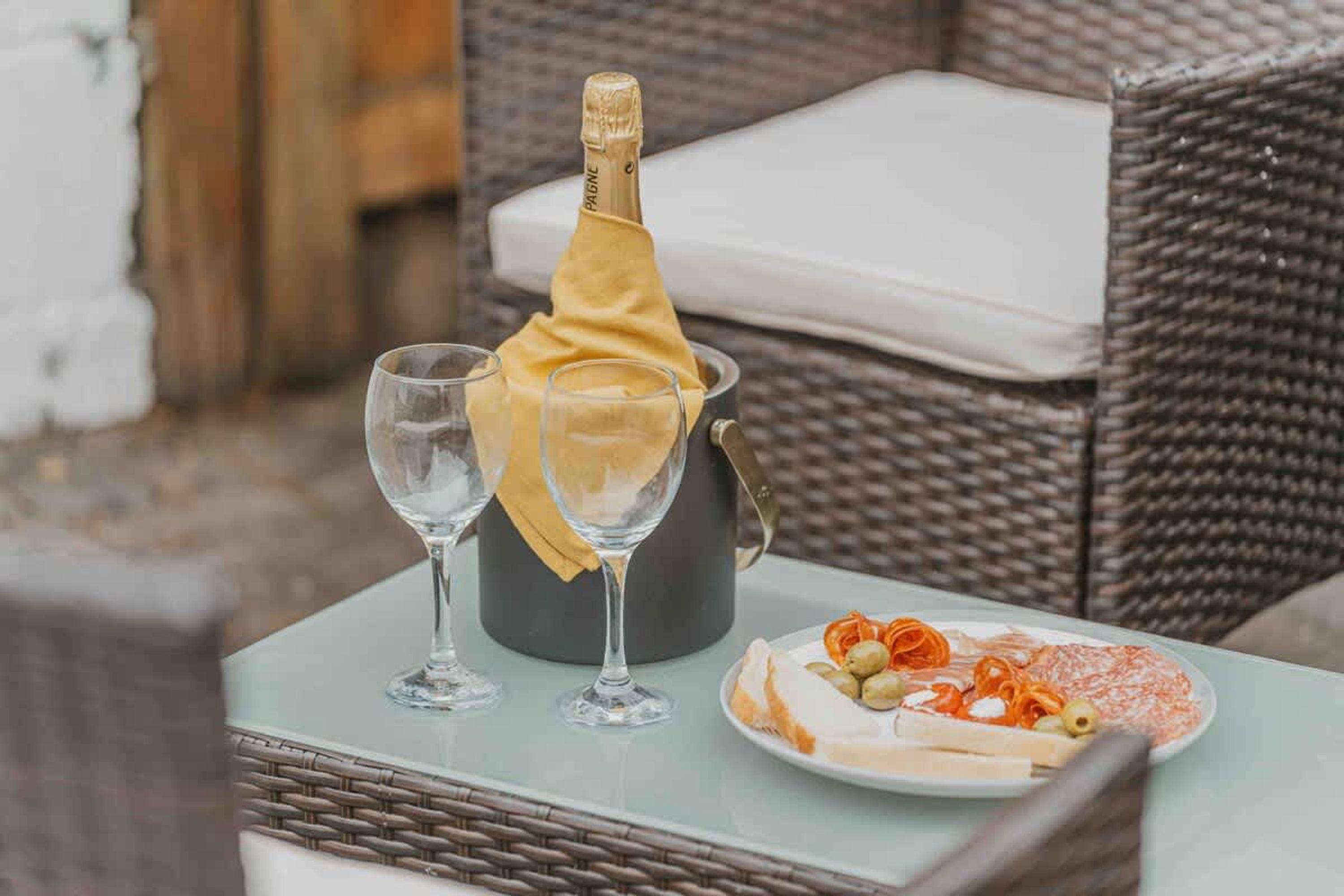 A wicker chair, a glass table, a bottle of champagne, and a plate with food.