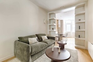 A living room with a green sofa, a round coffee table, and a white rug. There are shelves with decorative items and a window with curtains. - Luxury Duplex Saint-Germain Notre Dame (Paris)