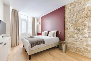 A bedroom with a bed, a TV, a stone wall, and a window with curtains. - Luxury Duplex Saint-Germain Notre Dame (Paris)