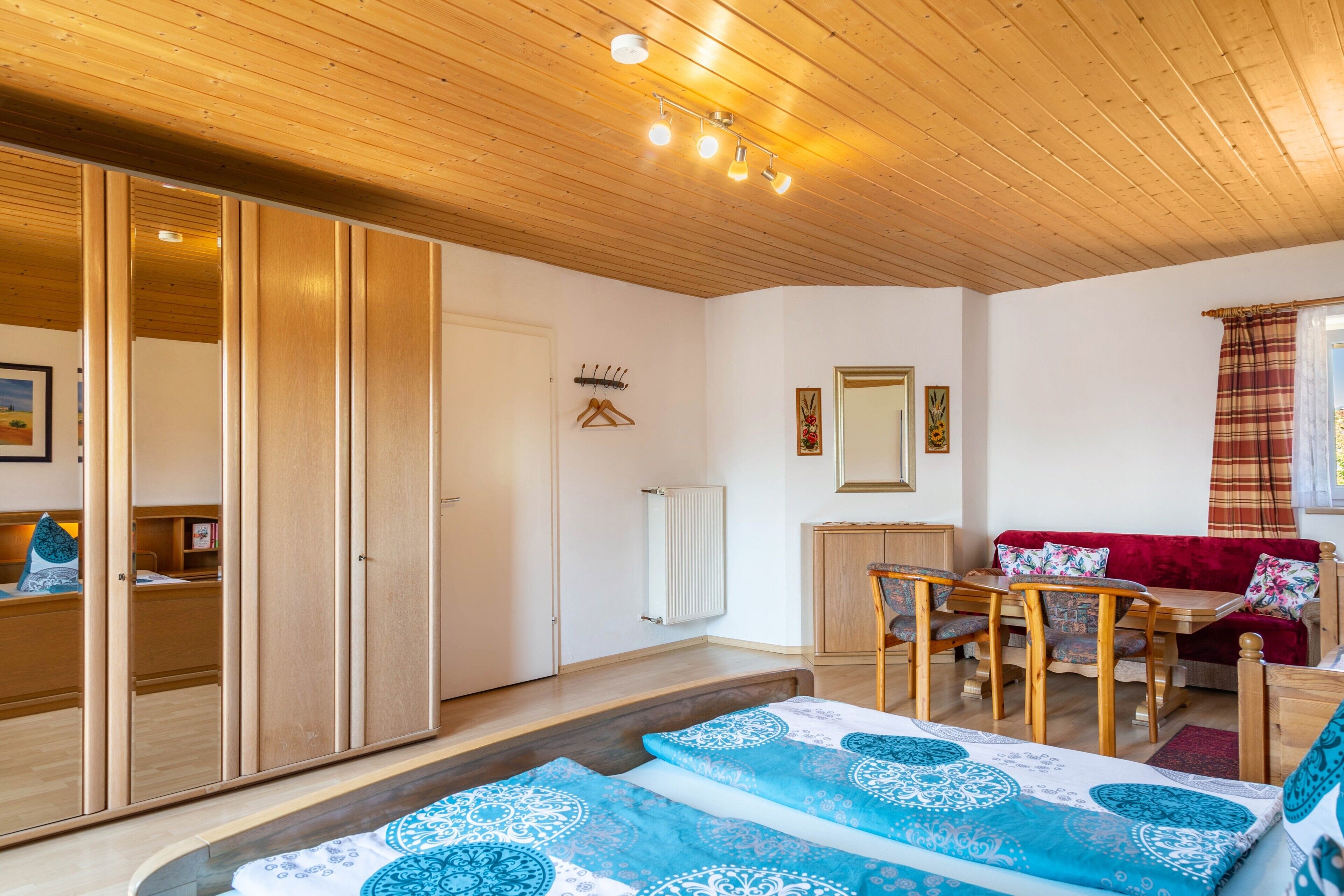 Ein gemütliches Zimmer mit Holzbalkendecke, einem Bett mit gemusterten Textilien, einem roten Sofa, einem Holztisch und einer Heizung.