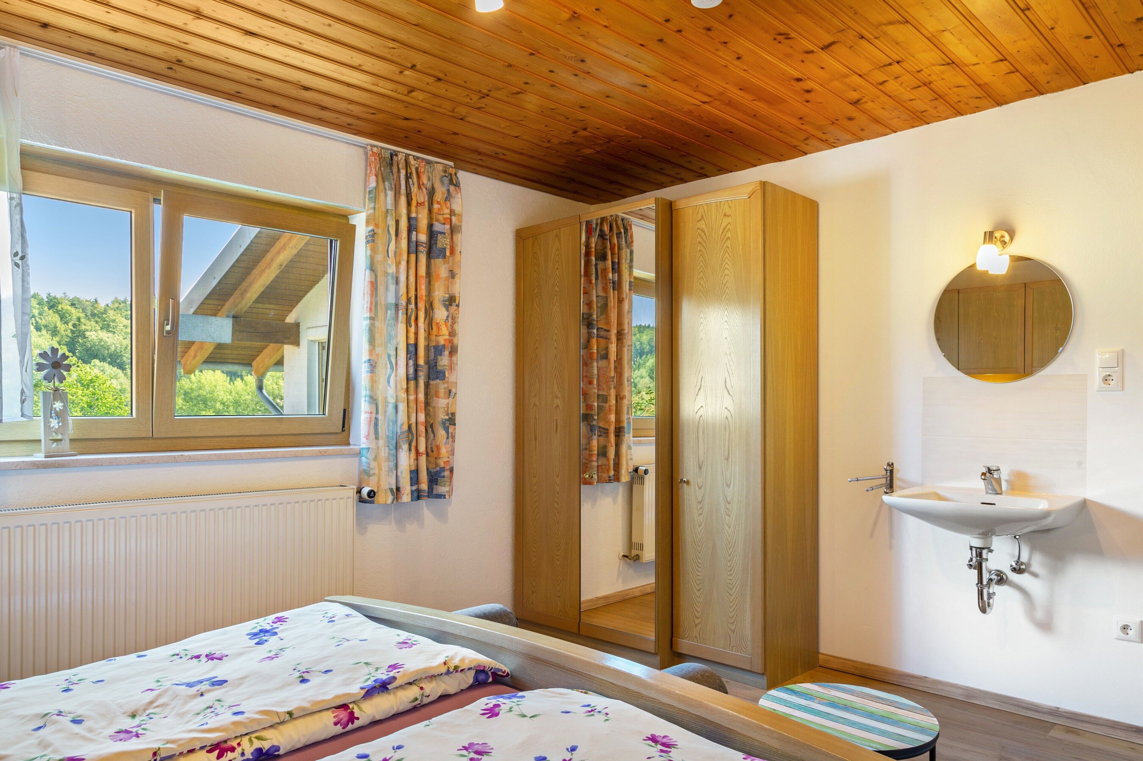 Ein Schlafzimmer mit einem Bett, einem Fenster, einem Badezimmer mit Waschbecken und Spiegel sowie einer Holzbalkendecke.