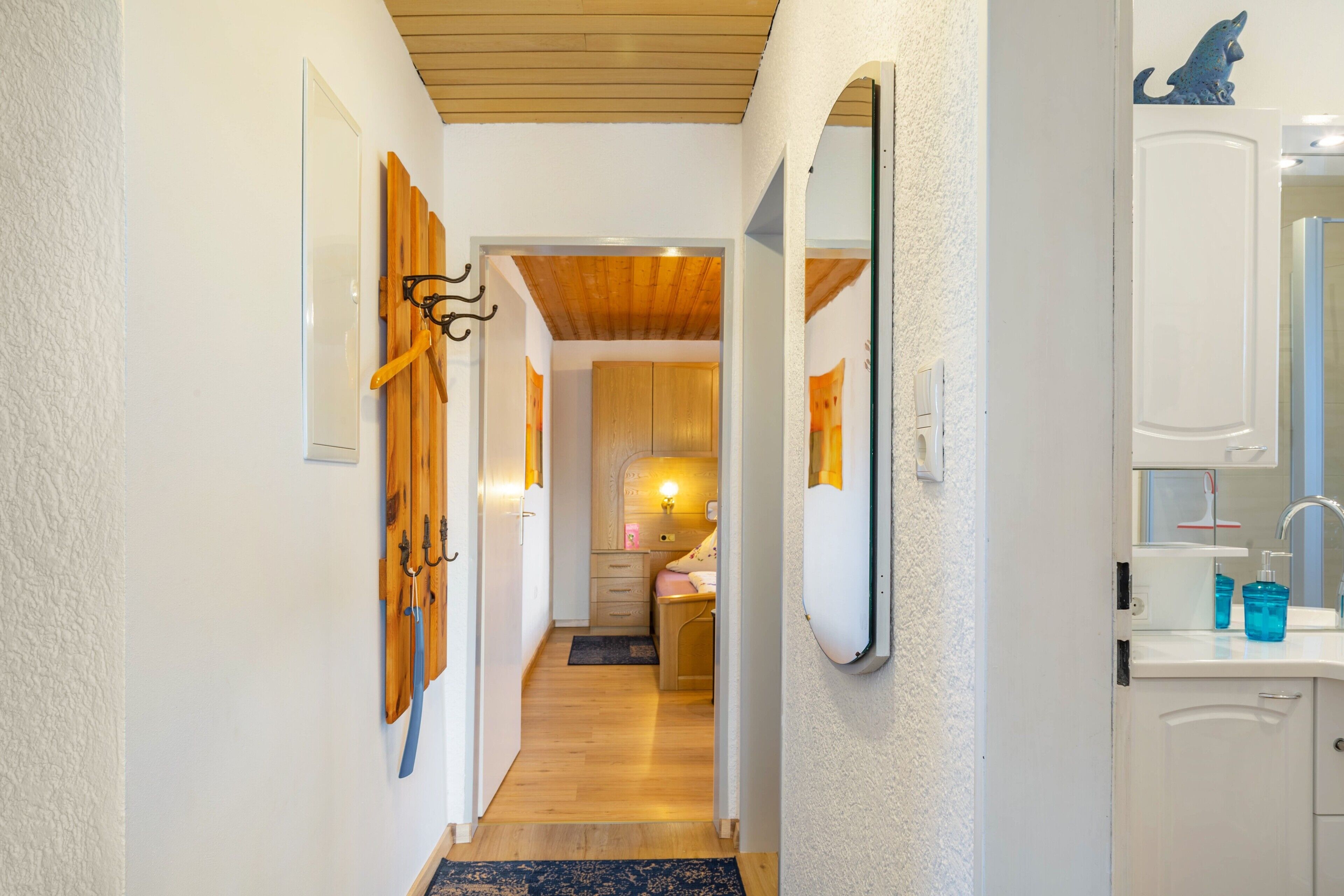 Un couloir avec des patères en bois fixées au mur, un miroir et une salle de bain équipée d’un lavabo et de produits de toilette.