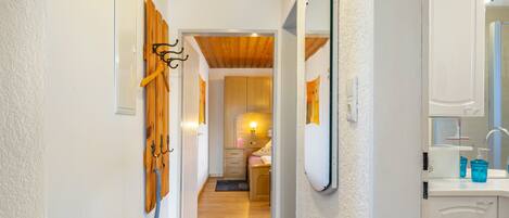 Un couloir avec des patères en bois fixées au mur, un miroir et une salle de bain équipée d’un lavabo et de produits de toilette.