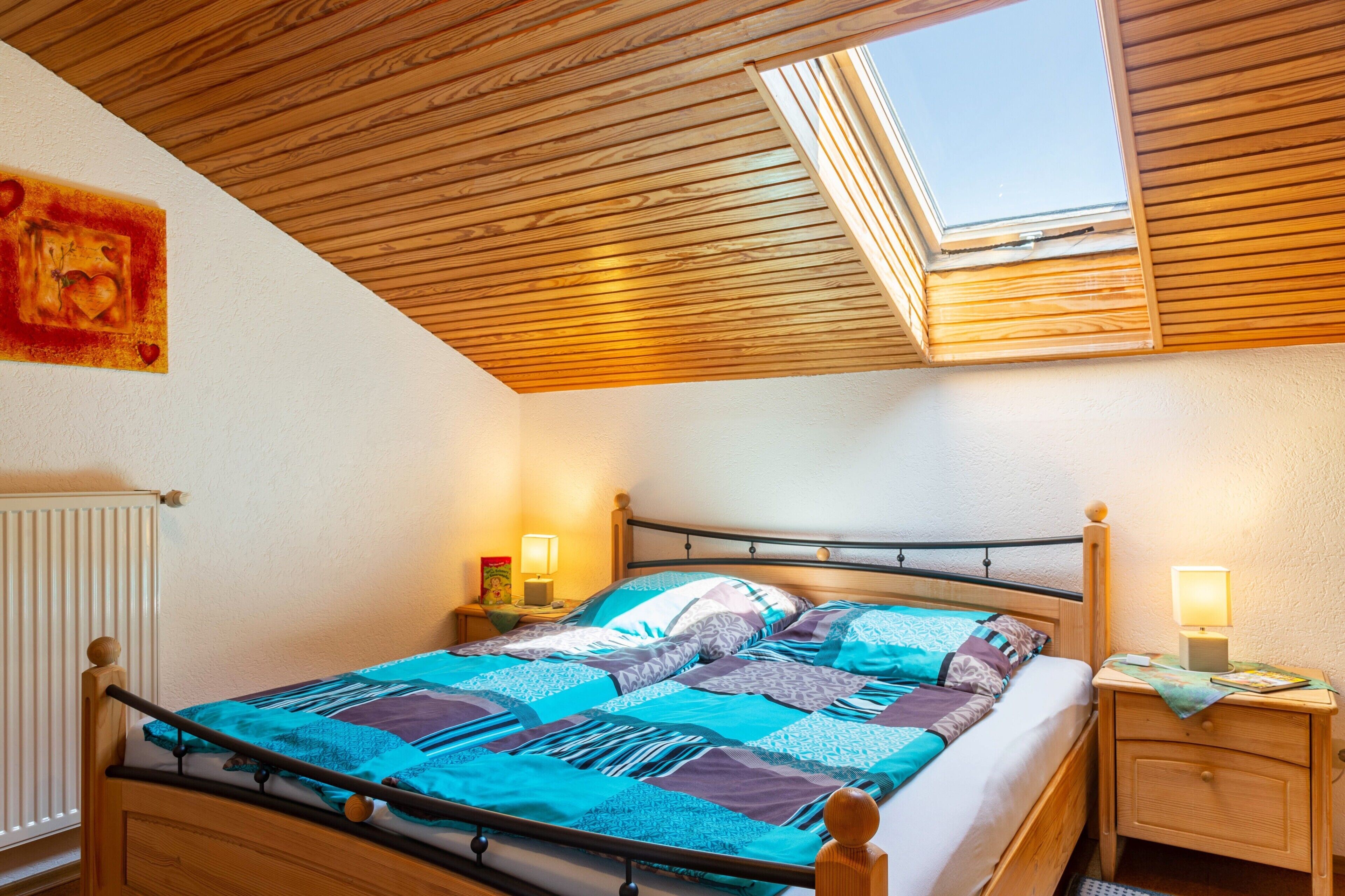 A bedroom with a sloped ceiling, a skylight, a bed with a wooden headboard, two bedside tables with lamps, and a radiator.