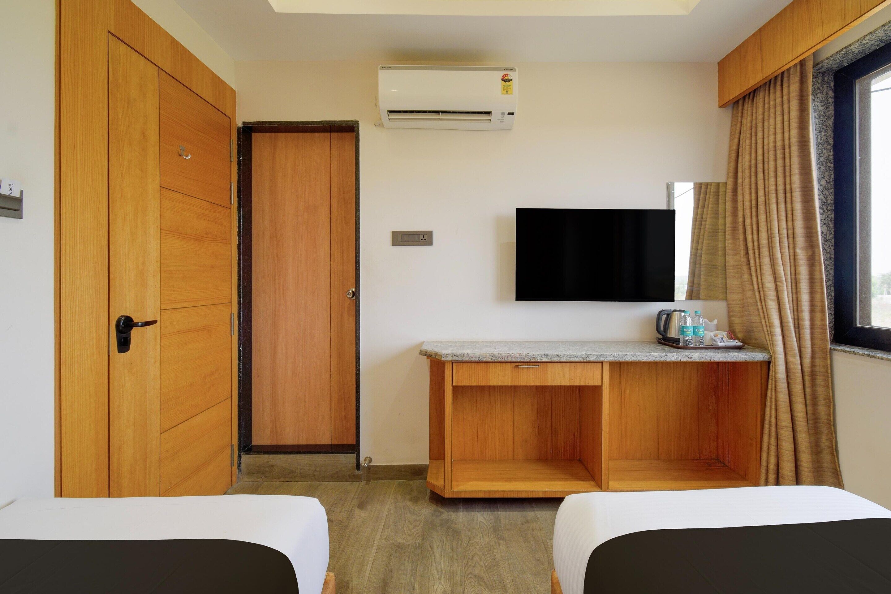 A hotel room with a wooden wardrobe, a desk with a flat-screen TV, a bed, and a window with curtains.
