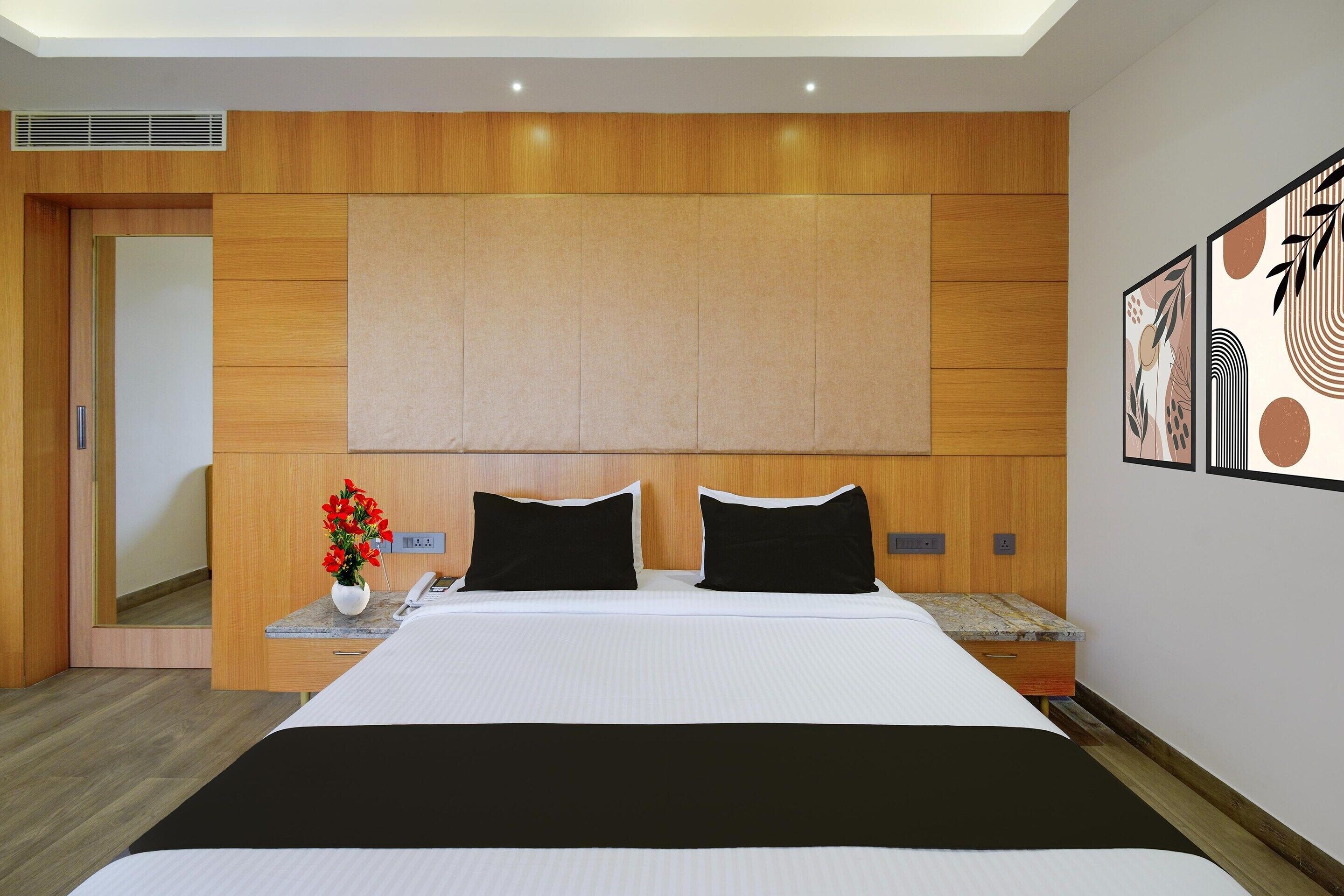 A modern hotel room with a large bed, wooden headboard, a nightstand with a vase of flowers, and a wall-mounted artwork.