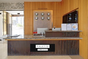 Un mostrador de recepción con un cartel que dice 'RECEPTION', un reloj con múltiples zonas horarias, una computadora y un cartel que dice 'PALETTE'.
