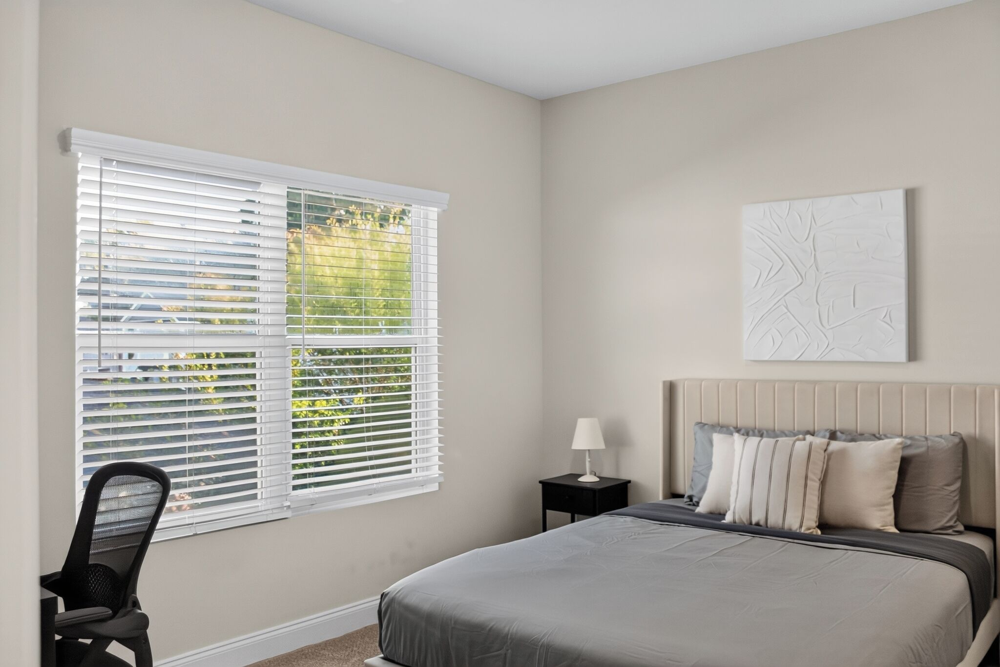 A bedroom with a bed, a nightstand, a chair, a window with blinds, and a wall-mounted artwork.