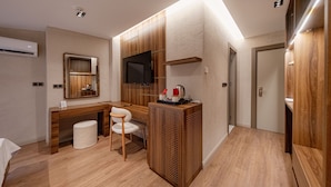 A modern hotel room with a wooden desk, a chair, a television, and a small kitchenette area.