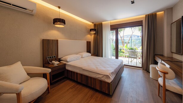 A modern hotel room with a wooden floor, a large bed, a desk, a chair, and a view of the outdoors.