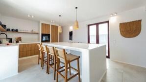 Una cocina moderna con una isla central, sillas de madera, luces colgantes y un ventanal grande.