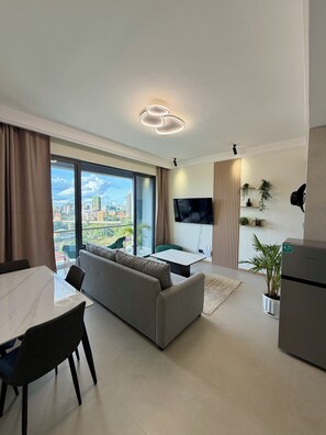 Een moderne woonkamer met een bank, eettafel en stoelen, een flatscreen televisie aan de muur en een groot raam met uitzicht over de stad.