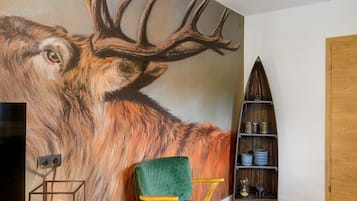 Ein Zimmer mit einer großen Wandmalerei eines Rehs, einem grünen Samt-Sessel, einem Holzregal und einem kleinen Tisch mit einer Lampe.