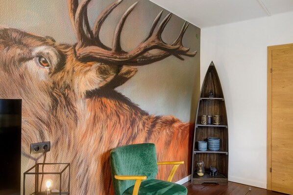 Una habitación con un amplio mural de un ciervo, un sillón de terciopelo verde, una estantería de madera y una mesita con una lámpara.