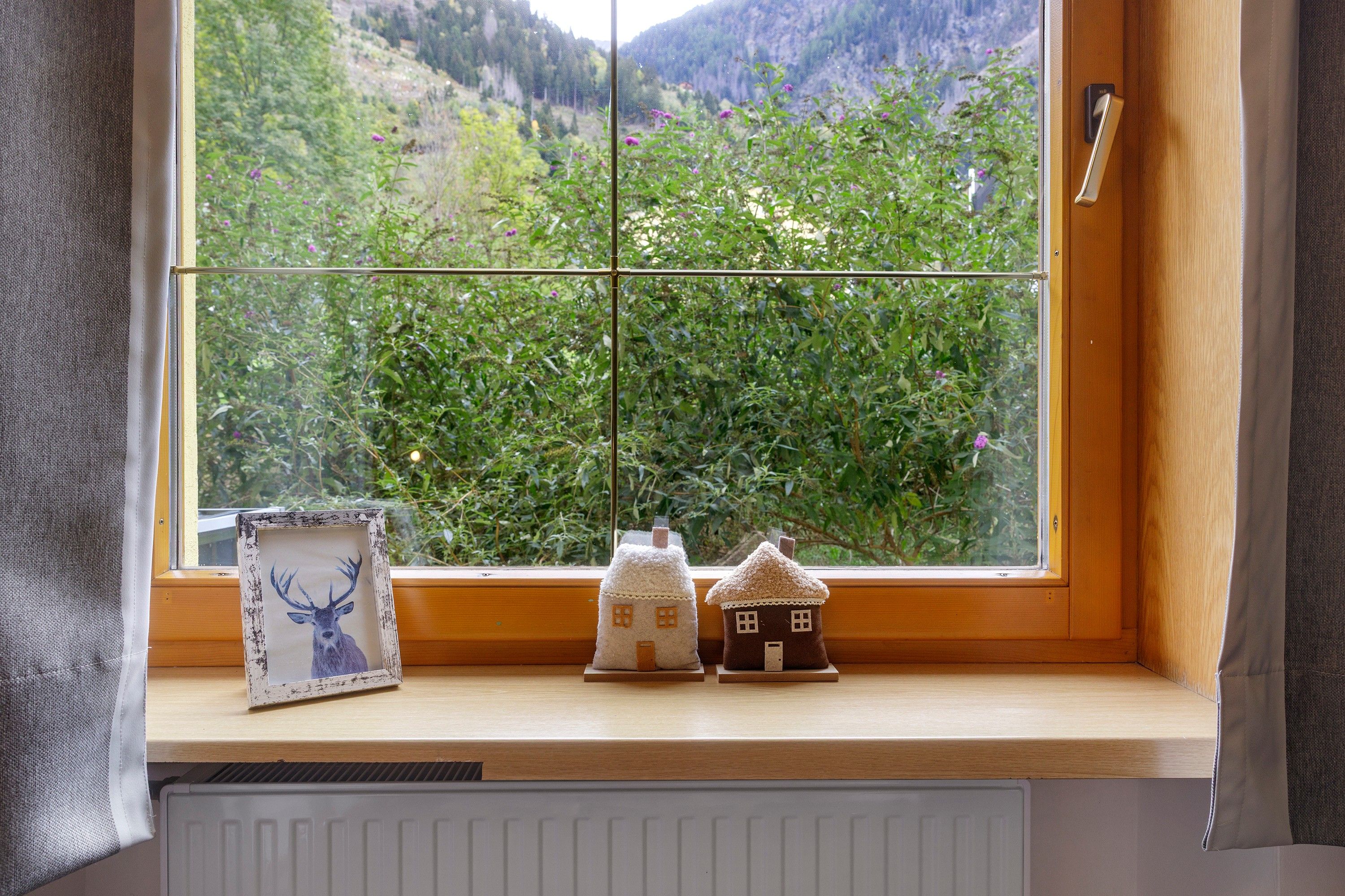 Una ventana con vista a un paisaje verde, un cuadro con un ciervo y dos casas pequeñas en el alféizar.