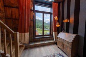Una habitación con escalera de madera, un ventanal con cortinas, un armario con una placa y una lámpara.