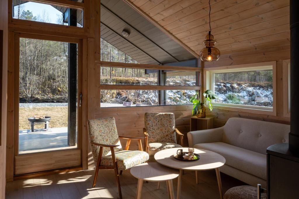 Een gezellig interieur van een blokhut met een bank, twee fauteuils, een ronde tafel en grote ramen met uitzicht op een bebost gebied.
