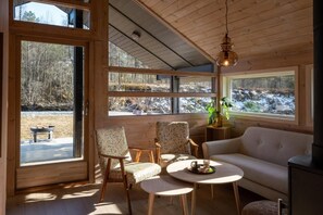 A cozy cabin interior with a sofa, two chairs, a round table, and large windows offering a view of a forested area.