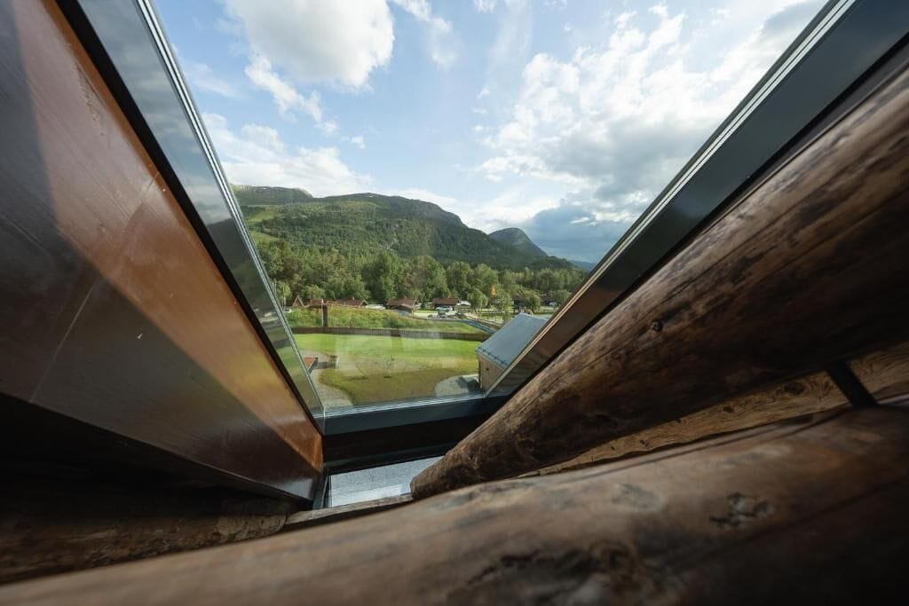 Vista a través de una ventana con marco de madera, que da a un paisaje verde con colinas y cielo despejado.