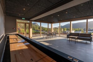 Un espacio interior amplio con mesa de billar, zona de estar con sofá y sillones, y grandes ventanales con vistas a un paisaje montañoso.