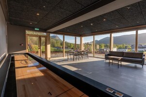 A spacious indoor area with a billiards table, a seating area with a sofa and chairs, and large windows offering a view of a mountainous landscape.