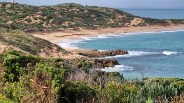 Un paisaje costero con un faro en una colina, playa arenosa y olas del océano.