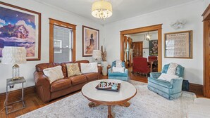 A living room with a brown leather sofa, a blue armchair, a white coffee table, and a chandelier. - Illini Dream House with Historic Retreat near UIUC and Downtown (Champaign)
