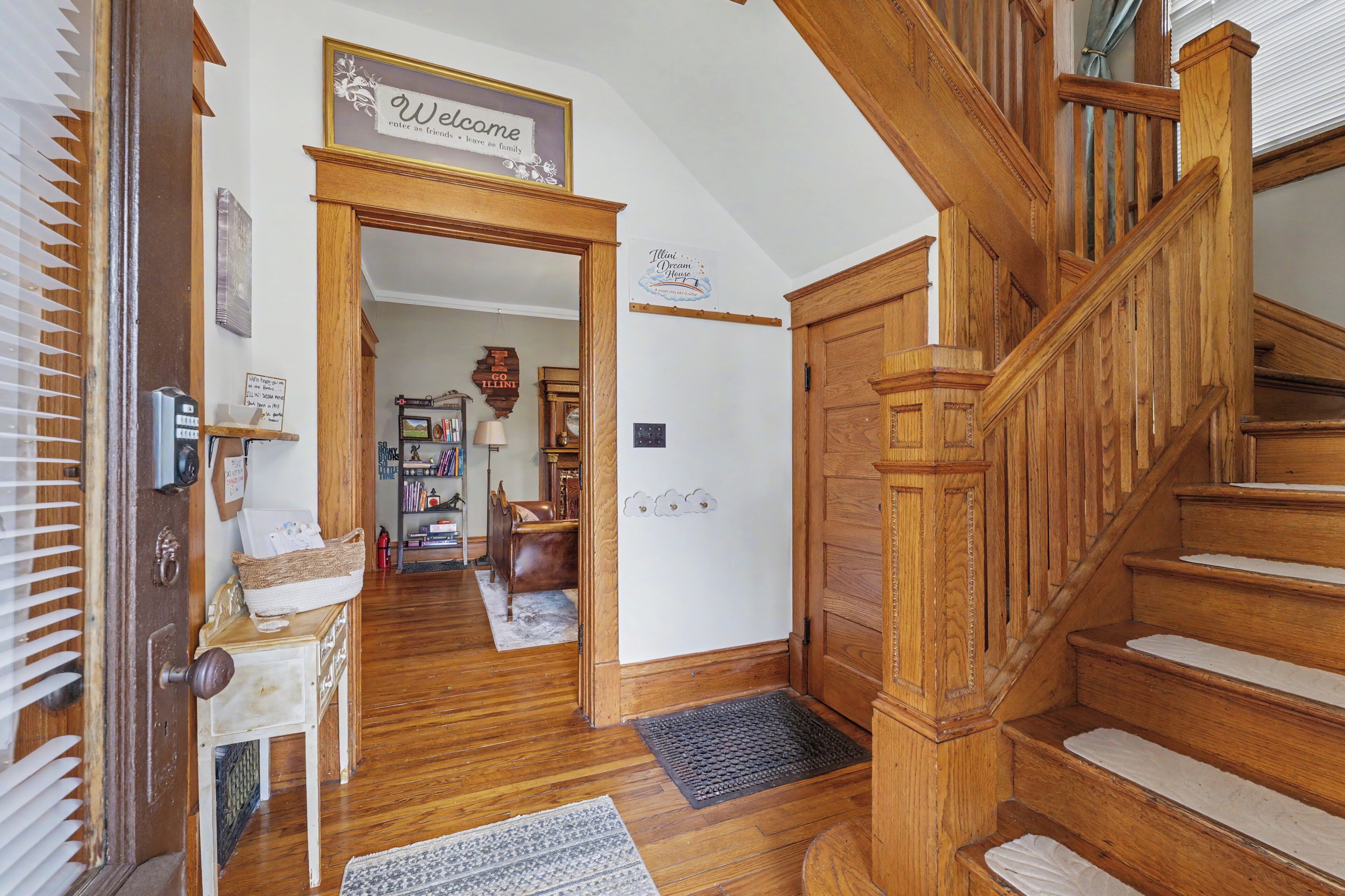 Un escalier en bois menant au deuxième étage, une pancarte de bienvenue, une porte avec un panier, et un couloir avec un tapis.