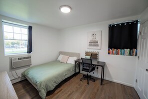 Une chambre à coucher avec un lit, un bureau avec une chaise, une fenêtre avec des rideaux, un climatiseur fixé au mur et une bibliothèque remplie de livres.