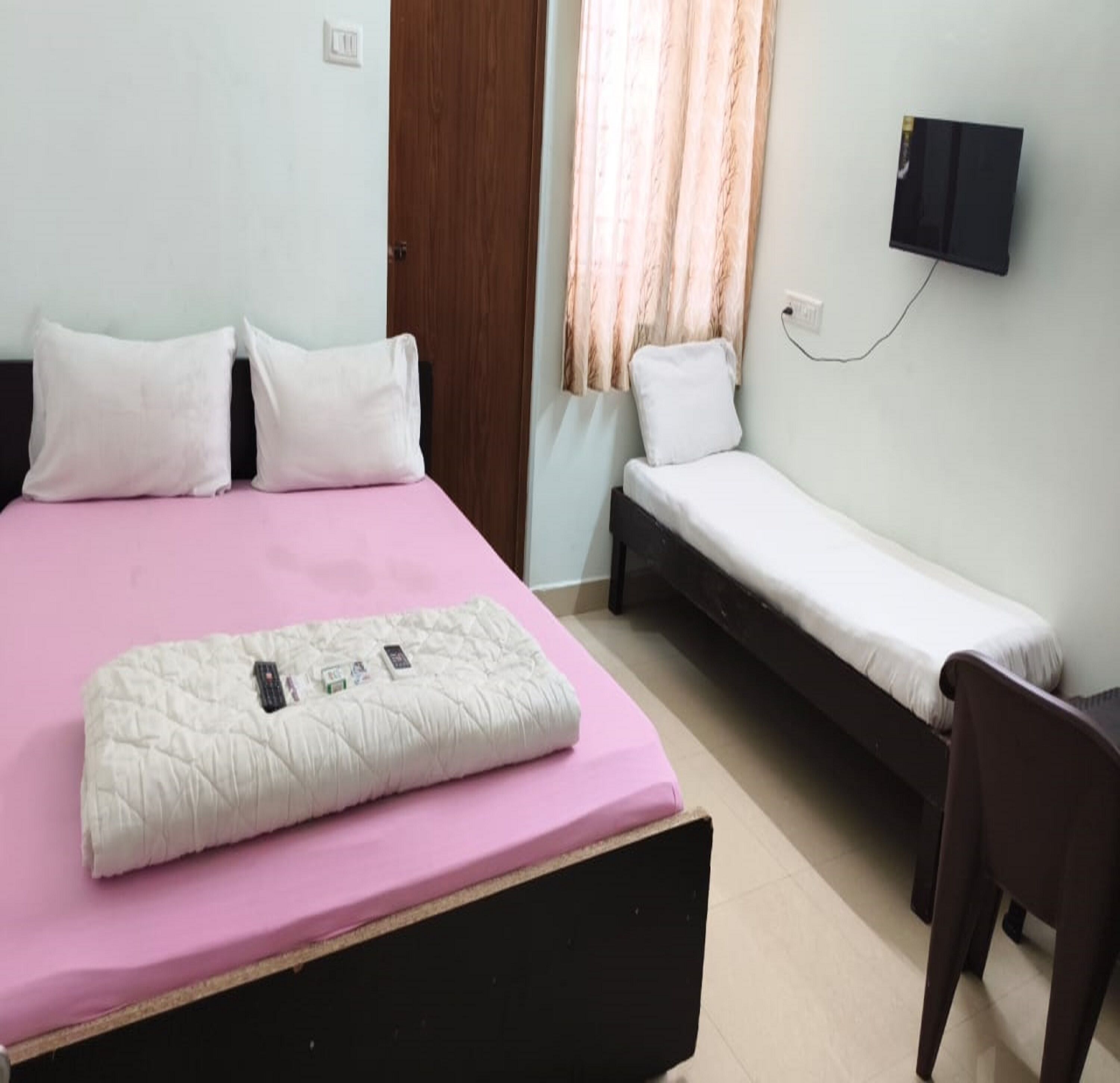 Une chambre à coucher avec un lit, une table de chevet, une chaise, un téléviseur et une fenêtre avec des rideaux.