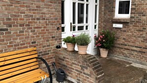 Een bakstenen muur met een houten bankje, een witte deur en plantenbakken.