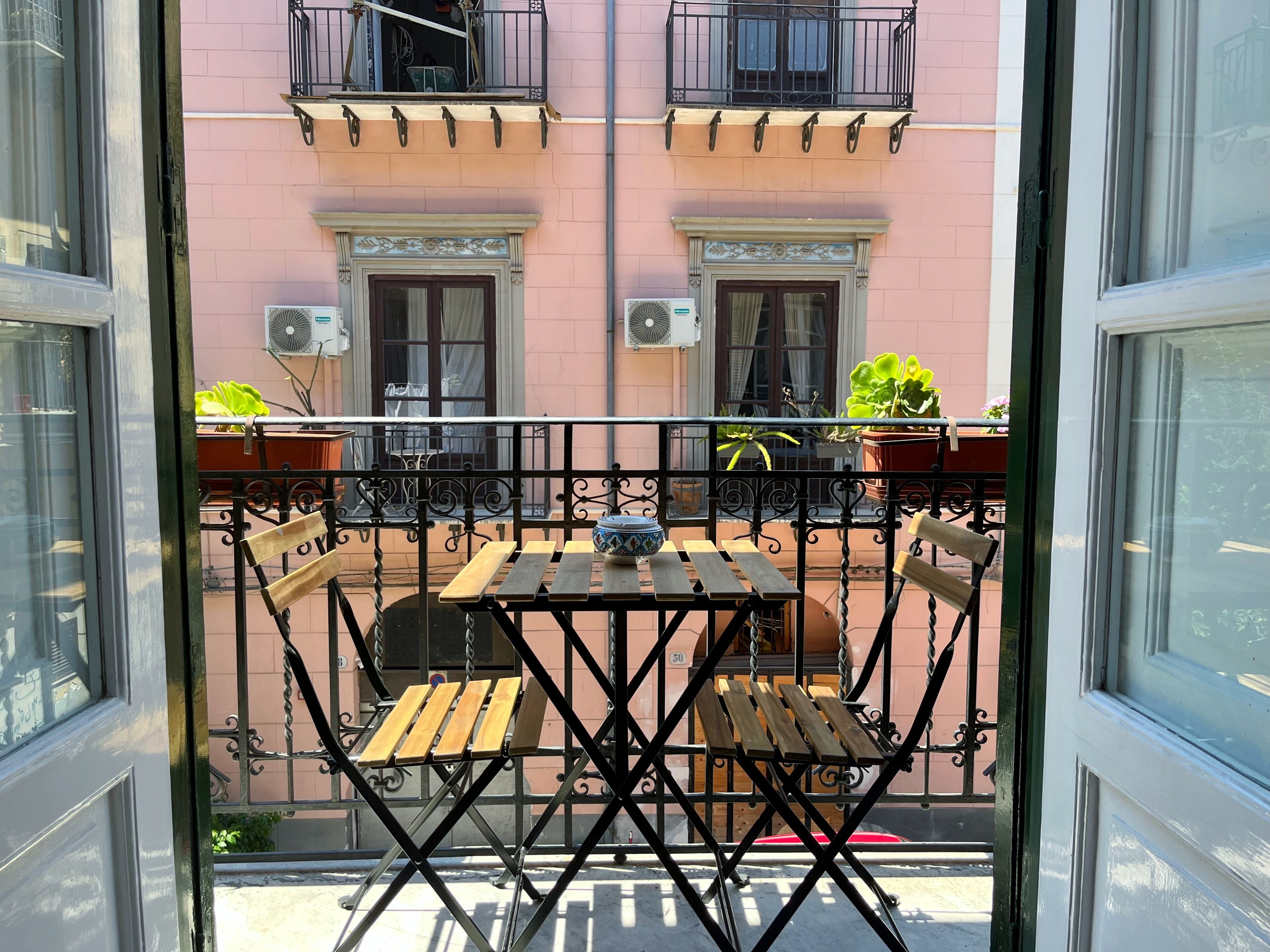 Ein Balkon mit Holztisch und -stühlen, einem Blumentopf und einem Fenster mit Blick auf ein rosafarbenes Gebäude.