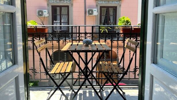 Un balcon avec une table et des chaises en bois, une plante en pot et une fenêtre donnant sur un bâtiment rose.
