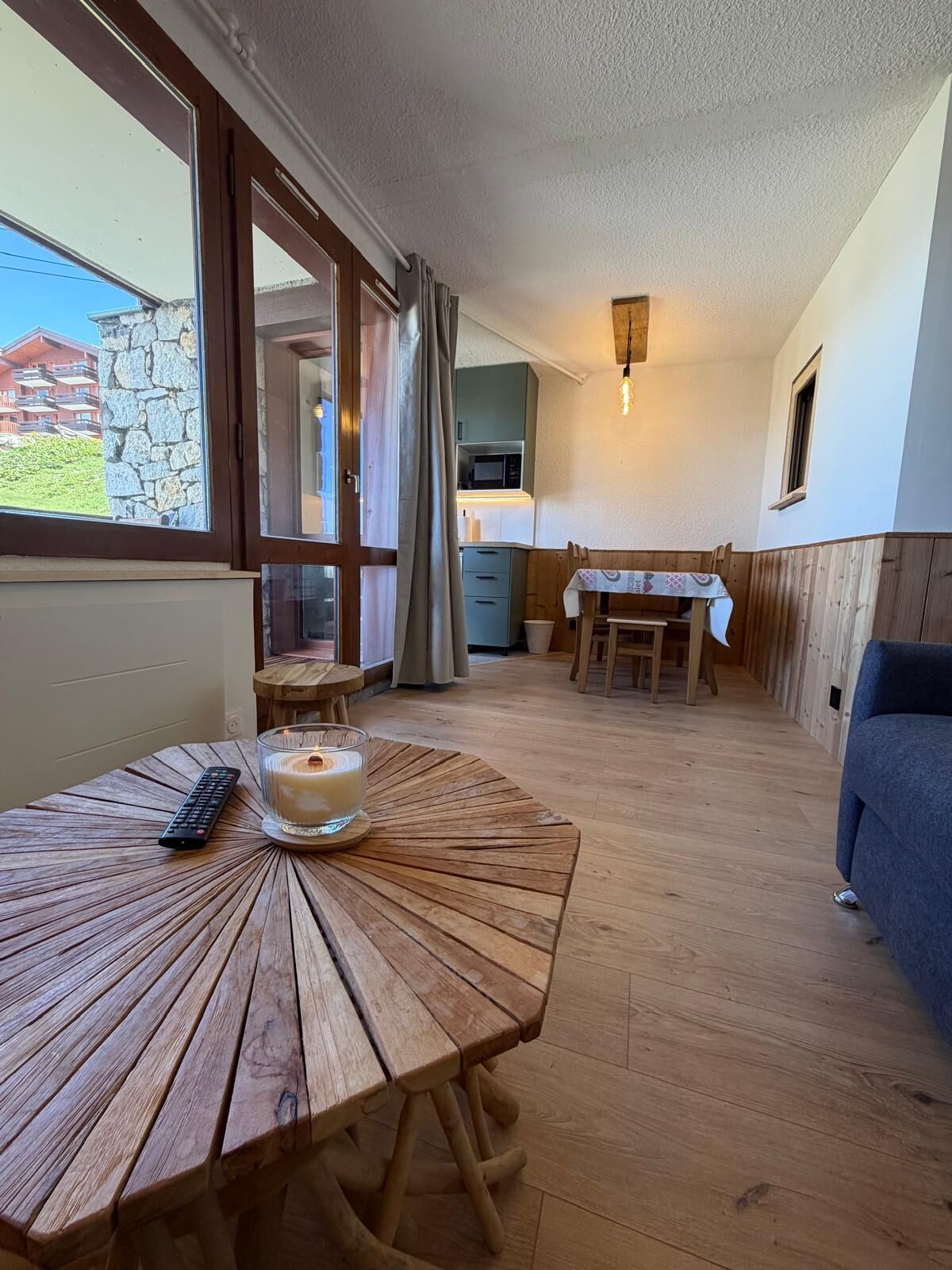 Une table en bois avec une télécommande et un verre de lait, un banc en bois, une table à manger avec des chaises, un canapé bleu et un mur en pierre avec une fenêtre.